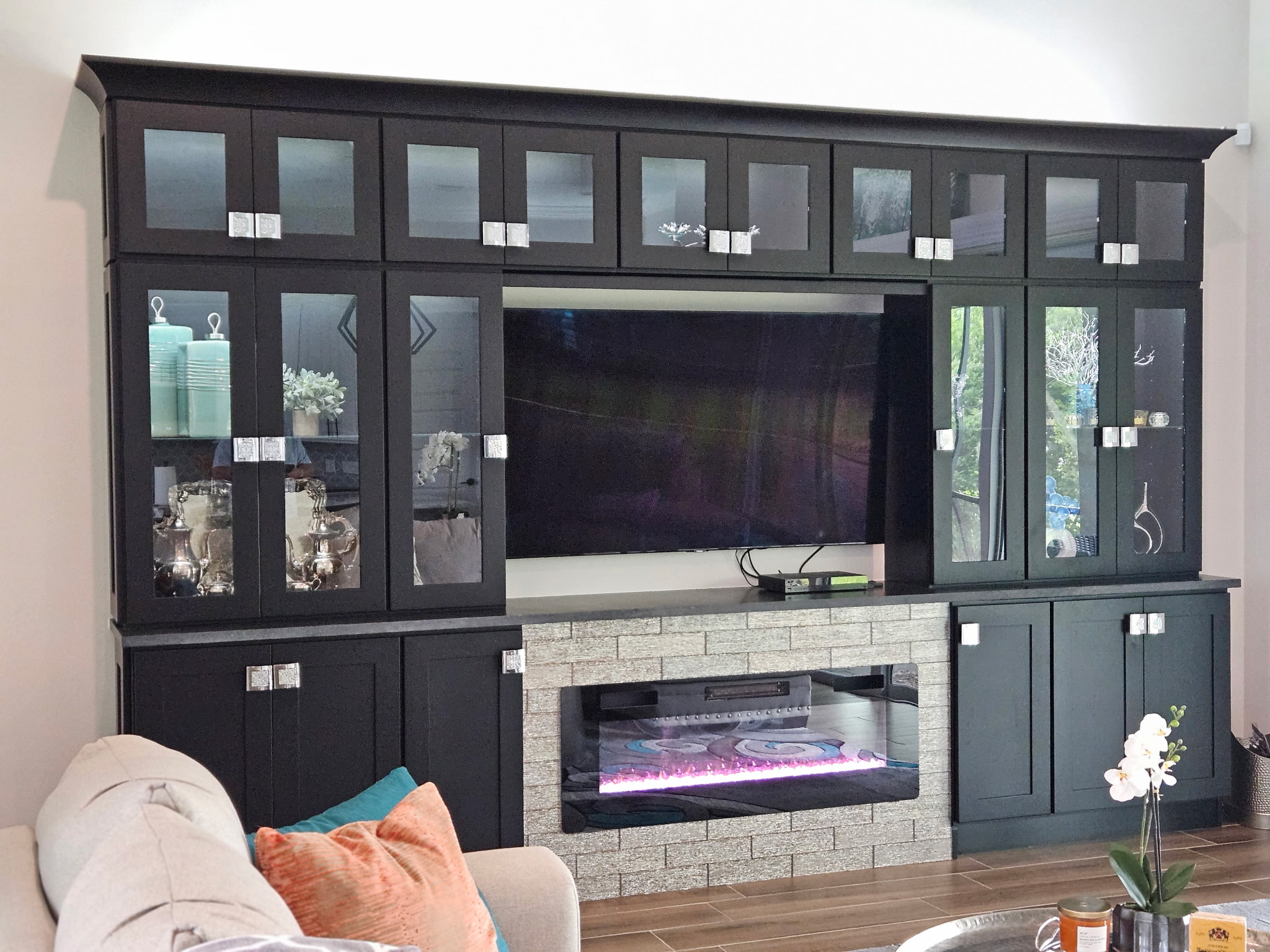 Black Shaker entertainment wall with glass-front upper cabinets, a centered TV, and an electric fireplace in a silver tile surround